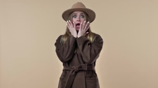 帽子とコートを身に着けている若い感情的な女性はスタジオでベージュの背景に隔離された何かの恐ろしい — ストック動画