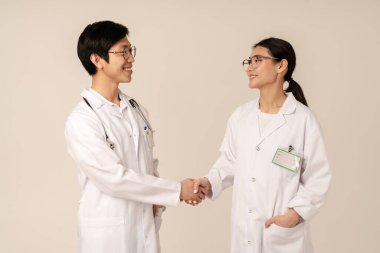 Beyaz üniformalı Asyalı genç tıp doktorlarının bej arka planda izole bir şekilde gülümseyip el sıkışmaları.