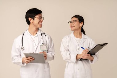 Beyaz üniformalı Asyalı genç tıp doktorlarının bej arka planda izole edilmiş, gülümseyen ve panoları tutan resimleri.