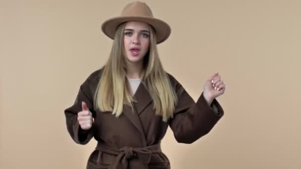 一个戴着帽子和外套的快乐的年轻女子在工作室的米色背景下独自跳舞 — 图库视频影像