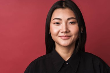 Kırmızı arka planda dikilip kameraya bakan, sıradan giyinmiş, güzel gülümseyen Asyalı genç bir kadının fotoğrafını çek.