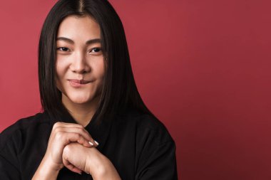 Kırmızı arka planda tek başına duran, sıradan kıyafetler giyen, gülümseyen genç Asyalı bir kadının fotoğrafını çek.