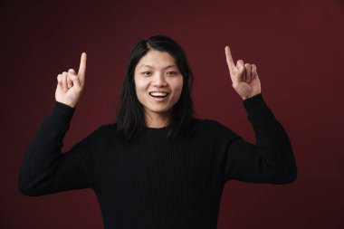 Karanlık arka planda izole edilmiş, günlük giysiler giymiş, fotokopi alanını işaret eden neşeli Asyalı genç bir kadının portresi.