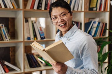 Kütüphanede durup kitap okuyan, gülümseyen, gözlüklü yakışıklı Asyalı bir adam resmi.