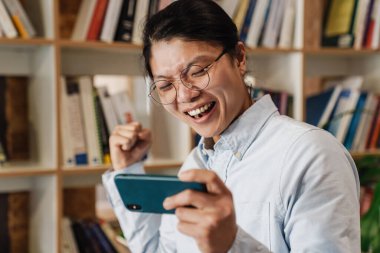 Cep telefonunda video oyunu oynayan heyecanlı Asyalı bir adamın görüntüsü ve kütüphanede ayakta dururken kazanan jest yapıyor