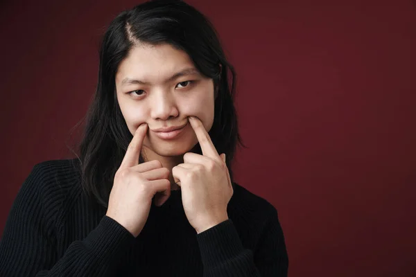 一个悲伤的亚洲男人用手指在勃艮第背景下制造假笑的图片 — 图库照片