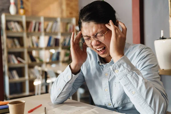 坐在餐桌前头痛 揉着太阳穴 尖叫的眼镜中的不快乐的亚洲人的形象 — 图库照片