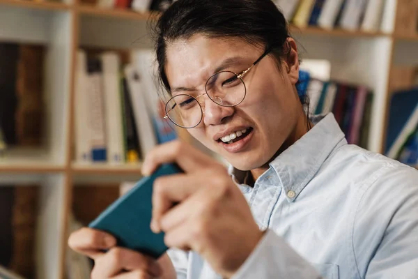 站在图书馆用眼镜在手机上玩电子游戏的不高兴的亚洲人的形象 — 图库照片