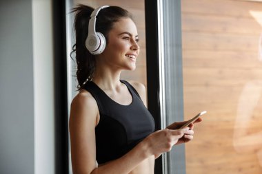 Spor kıyafetleri içinde gülümseyen baştan çıkarıcı bir kadının pencerenin yanında dururken kulaklık ve akıllı telefon kullandığı fotoğraf.