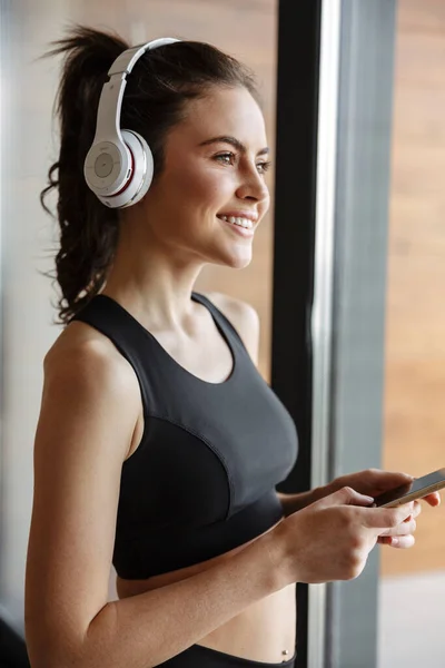 Zdjęcie Uśmiechniętej Uwodzicielki Odzieży Sportowej Używającej Słuchawek Smartfona Stojącej Przy — Zdjęcie stockowe