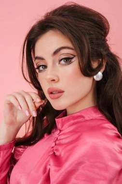 Parlak makyajlı güzel bir esmer kadının portresi şık kıyafetler giyiyor pembe arka planda izole bir şekilde kameraya bakıyor.