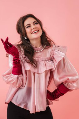 Parlak makyajlı, güzel, genç, esmer bir kadının portresi. Pembe arka planda tek başına duran şık kıyafetler giyiyor.