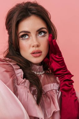 Parlak makyajlı, şık giyinen, pembe arka planda tek başına duran güzel esmer bir kadının portresi.