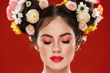 Güzel, genç, esmer bir kadının portresi. Çiçekli saç stili ve parlak makyajlı. Kırmızı arka planda izole edilmiş poz veriyor.