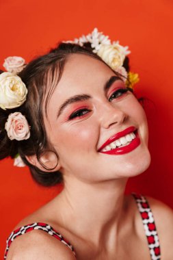Güzel, gülümseyen bir esmer kadının portresi. Çiçekli saç stili ve parlak makyajlı. Kırmızı arka planda izole edilmiş poz veriyor.