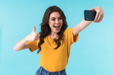 Duygusal olarak mutlu, iyimser, güzel bir kadının mavi duvar arkasında izole edilmiş bir şekilde poz vermesi. Cep telefonuyla selfie çekin ve onaylayın..