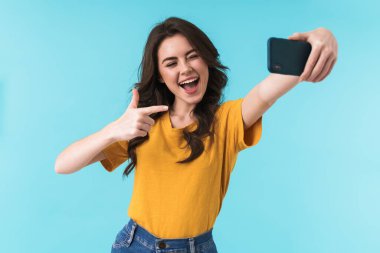 Duygusal olarak mutlu, iyimser, güzel bir kadının görüntüsü. Mavi duvar arkasında izole bir şekilde poz veriyor. Cep telefonuyla selfie çekiyor..