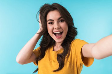 Duygusal olarak mutlu, neşeli, genç ve güzel bir kadının görüntüsü. Mavi duvar arkasında izole bir şekilde poz veriyor. Kamerayla selfie çek..