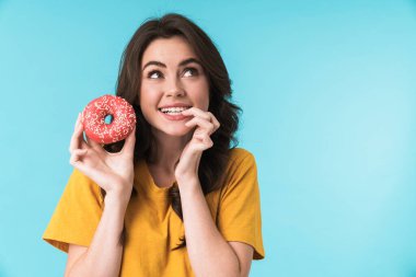 Mavi duvar arkasında izole edilmiş poz veren güzel bir kadın hayal etmek. Elinde donutla yan tarafa bakıyor..