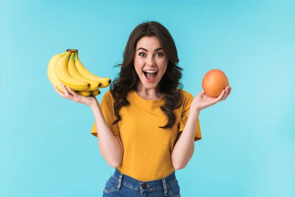 照片上的年轻漂亮女子被隔离在蓝色的墙后面 手里拿着香蕉和橙子 高兴极了 — 图库照片