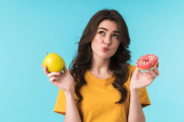 想象一下 一个心事重重的年轻貌美的女人站在蓝色的墙后面 手里拿着苹果和甜甜圈 — 图库照片