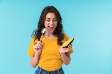 Avokado ve kaşık tutarak mavi duvar arkasında poz veren tatlı, heyecanlı, mutlu bir kadının fotoğrafı..