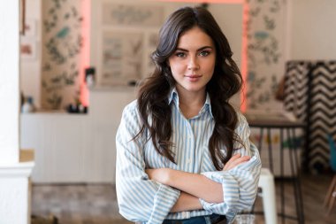 Kapalı bir kafede kollarını kavuşturmuş, resmi tişört giymiş, gülümseyen esmer bir kadının fotoğrafını çek.