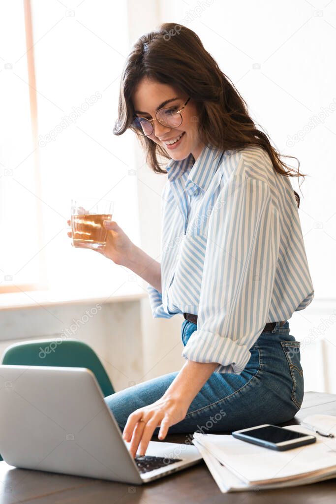 Attractive smiling young brunette woman studying in the cafe indoors, working on laptop computer, sitting on a table