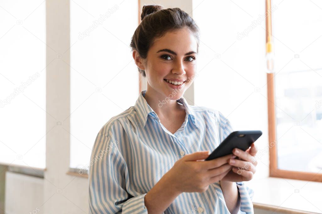 Attractive smiling young brunette businesswoman wearing formal shirt standing at the window indoors, using mobile phone