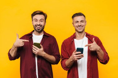 Kırmızı tişörtlü, gülümseyen ve sarı arka planda izole edilmiş cep telefonlarını işaret eden iki yakışıklı adam.