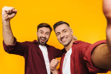 İki çekici, neşeli genç erkek arkadaş günlük kıyafetler giyip sarı arka planda izole bir şekilde selfie çekiyorlar.