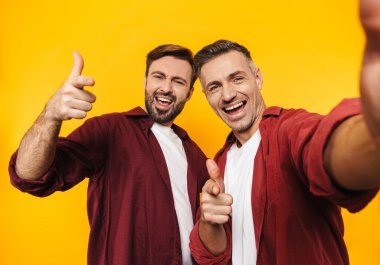 İki çekici, neşeli genç erkek arkadaş günlük kıyafetler giyip sarı arka planda izole bir şekilde selfie çekiyorlar.