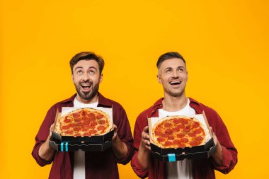 Sarı arka planda izole bir şekilde duran, kutuda pizza gösteren, gündelik kıyafetler giyen iki çekici, neşeli genç erkek arkadaş.