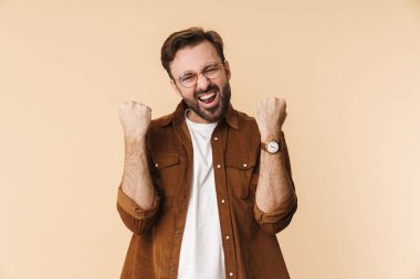 Bej arka planda izole edilmiş günlük kıyafetler giyen neşeli, genç sakallı bir adamın portresi başarıyı kutluyor.