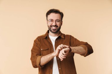 Neşeli, genç, sakallı bir adamın portresi. Günlük giysiler giymiş. Bej arka planda tek başına duruyor.