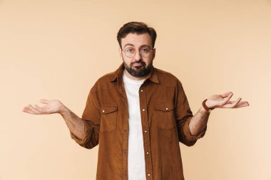 Kafası karışmış genç sakallı bir adamın portresi. Günlük kıyafetler giymiş. Bej arka planda tek başına duruyor, omuzlarını silkiyor.