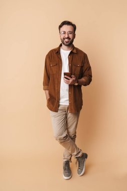 Neşeli, sakallı, sıradan kıyafetler giyen, bej arka planda tek başına duran, elinde cep telefonu olan genç bir adamın uzun bir portresi.