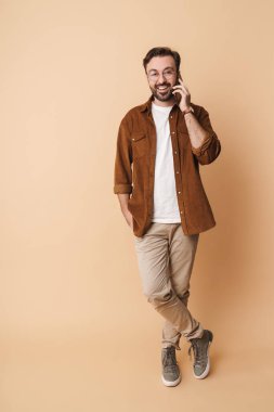 Neşeli, sakallı, sıradan kıyafetler giyen, bej arka planda izole bir şekilde cep telefonuyla konuşan genç bir adamın tam boy portresi.