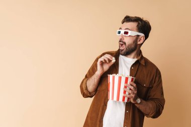 Neşeli, genç sakallı bir adamın portresi. Günlük giysiler giyip bej arka planda tek başına dikiliyor. 3D gözlüklü bir film izliyor, patlamış mısır yiyor, gözlerini kaçırıyor.