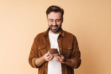 Neşeli, genç, sakallı bir adamın portresi. Günlük kıyafetler giymiş, bej arka planda izole bir şekilde duruyor. Elinde doğum günü pastasından bir parça tutuyor.