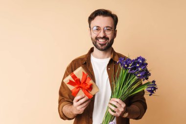 Bej arka planda izole bir şekilde duran, elinde bir buket çiçek ve hediye kutusuyla günlük giysiler giyen neşeli, genç sakallı bir adamın portresi.