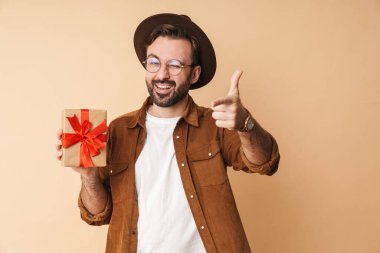 Şapkalı, mutlu, iyimser genç bir adamın bej rengi arka plan üzerinde izole edilmiş ve elinde sana işaret eden bir hediye kutusu..