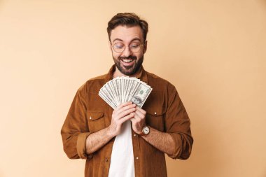 Tıraşsız, mutlu, genç bir adamın bej duvar arkasında izole edilmiş ve elinde para tutan görüntüsü..