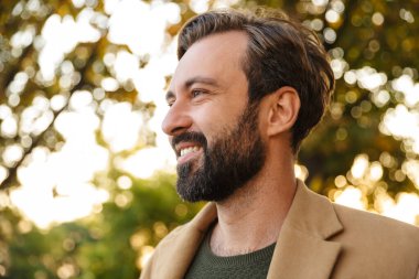 Ceketli, yakışıklı, beyaz sakallı ve parkta gülümseyen yetişkin bir adam.