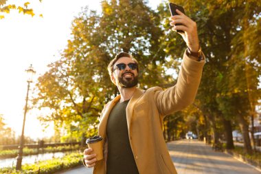 Ceketli yakışıklı yetişkin bir adamın fotoğrafı. Parkta yürürken gülümseyen ve fotoğraf çeken.