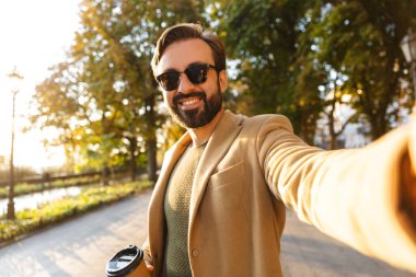 Ceketli yakışıklı yetişkin bir adamın fotoğrafı. Parkta yürürken gülümseyen ve fotoğraf çeken.