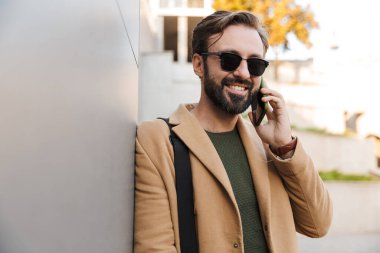 Güneş gözlüklü, yakışıklı, beyaz sakallı yetişkin bir adamın resmi. Akıllı telefondan konuşuyor ve binanın yanında dikiliyor.