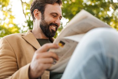 Ceketli, yakışıklı, yetişkin bir adamın parkta bankta otururken gülümseyip gazete okuduğu bir görüntü.