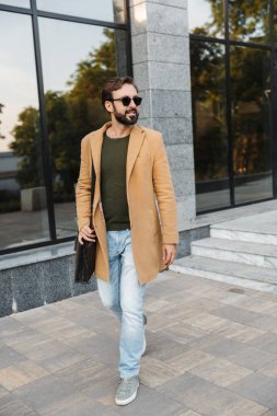 Kafkasyalı yakışıklı, güneş gözlüklü yetişkin bir adam. Şehir caddesinde modern binanın yanında yürüyor.