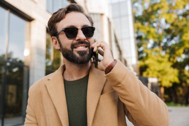 Gözlüklü, yakışıklı, sakallı, yetişkin bir adamın şehir caddesinde yürürken telefonla konuşması.